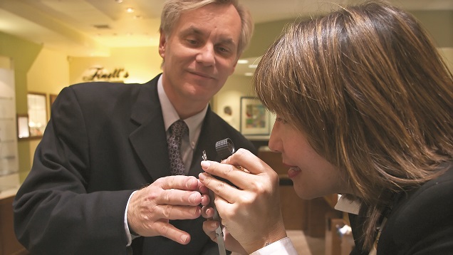 A male retail jeweler helps a woman look at a diamond through a loupe.